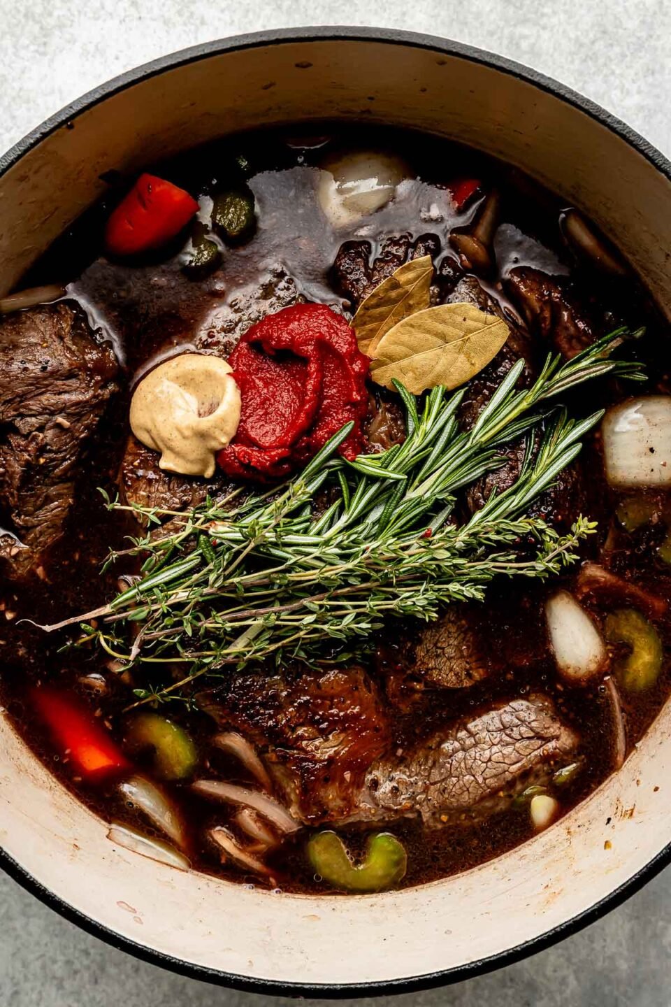 An overhead shot of beef chuck braising with fresh herbs, vegetables, dijon mustard, bay leaves, tomato paste, red wine and beef stock in a Dutch oven atop a light grey surface.