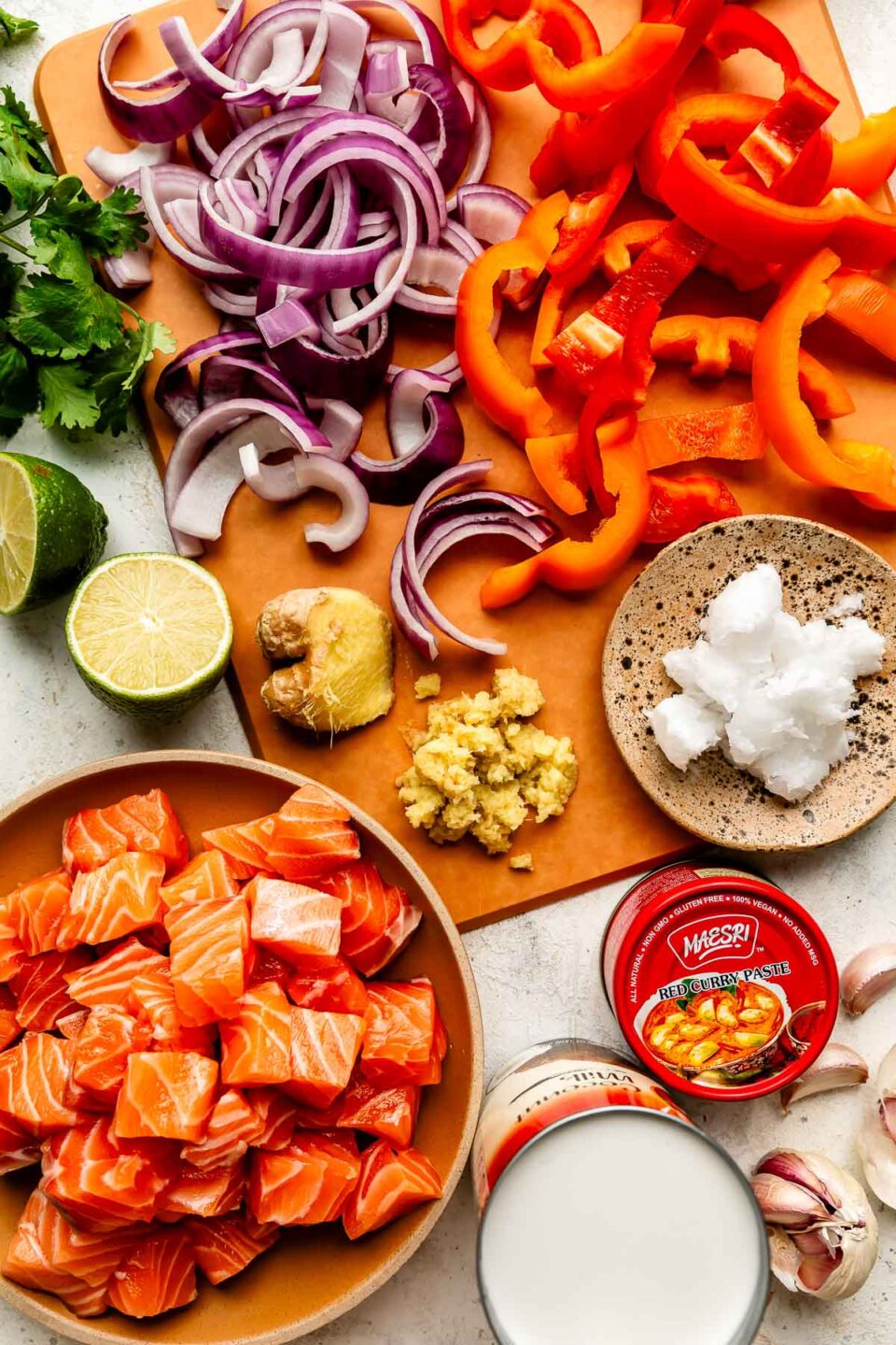 An overhead shot of ingredients displayed on a brown cutting board and a white marbled surface: sliced red pepper and red onion, lime, raw salmon cubes, garlic, ginger, cilantro, coconut milk, and red curry paste.