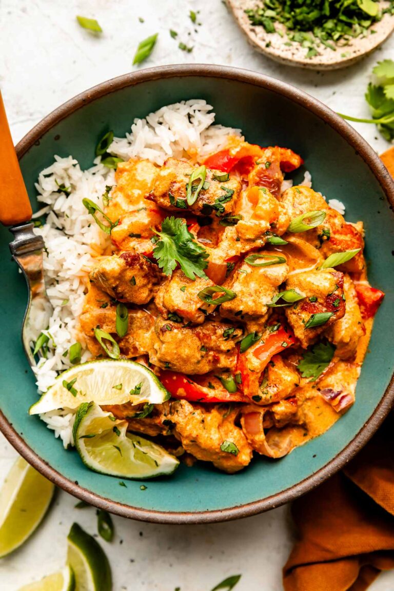 An overhead shot of a green bowl of salmon red curry served over white rice. The bowl sits on a white surface alongside dishes of cilantro and green onion, an orange cloth, and several lime wedges.