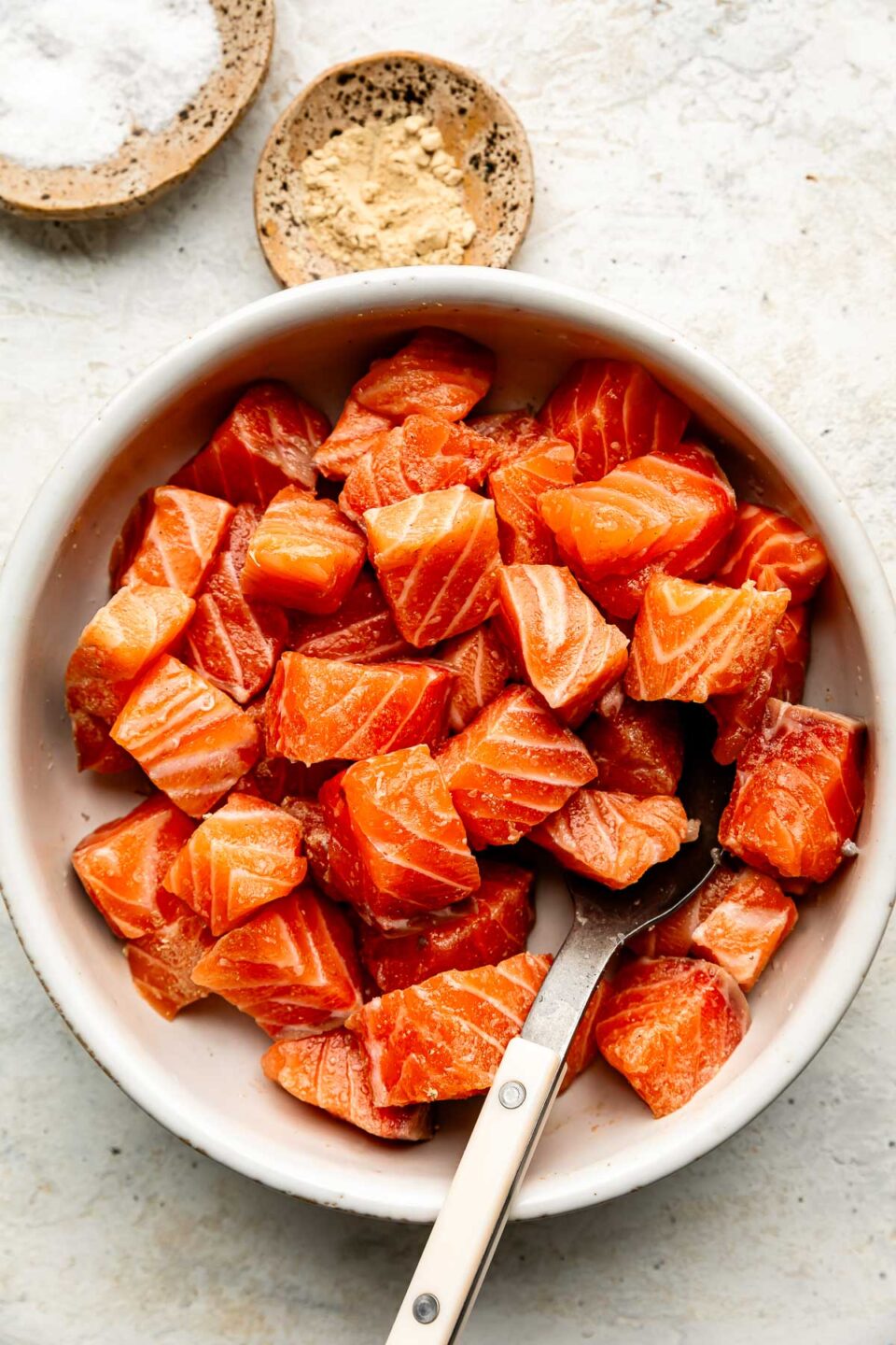 An overhead shot of raw cubed salmon in a white bowl atop an off-white textured surface. Dishes of salt and pepper sit alongside the bowl.