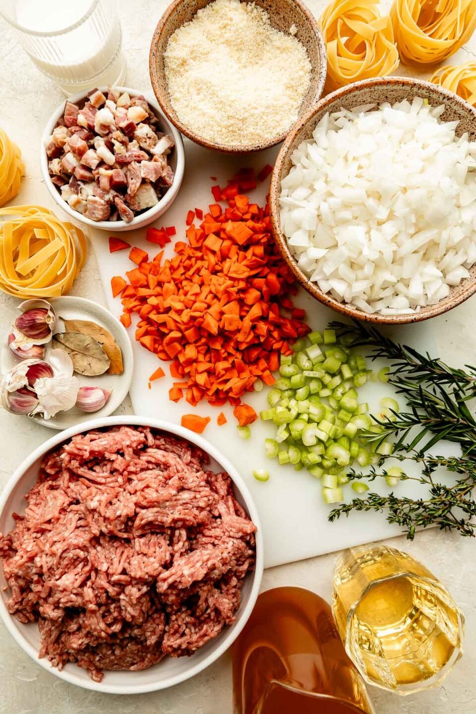 Ingredients are displayed on a white cutting board and off-white surface: chopped carrots, celery, and onions, ground pork, garlic, white wine, dry pasta, grated parmesan, pancetta, broth, heavy cream and fresh herbs.