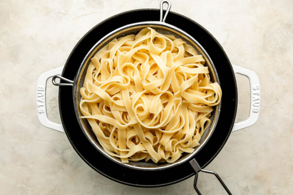 An overhead shot of cooked tagliatelle noodles in a silver colander over a white Dutch oven atop an off-white textured surface.