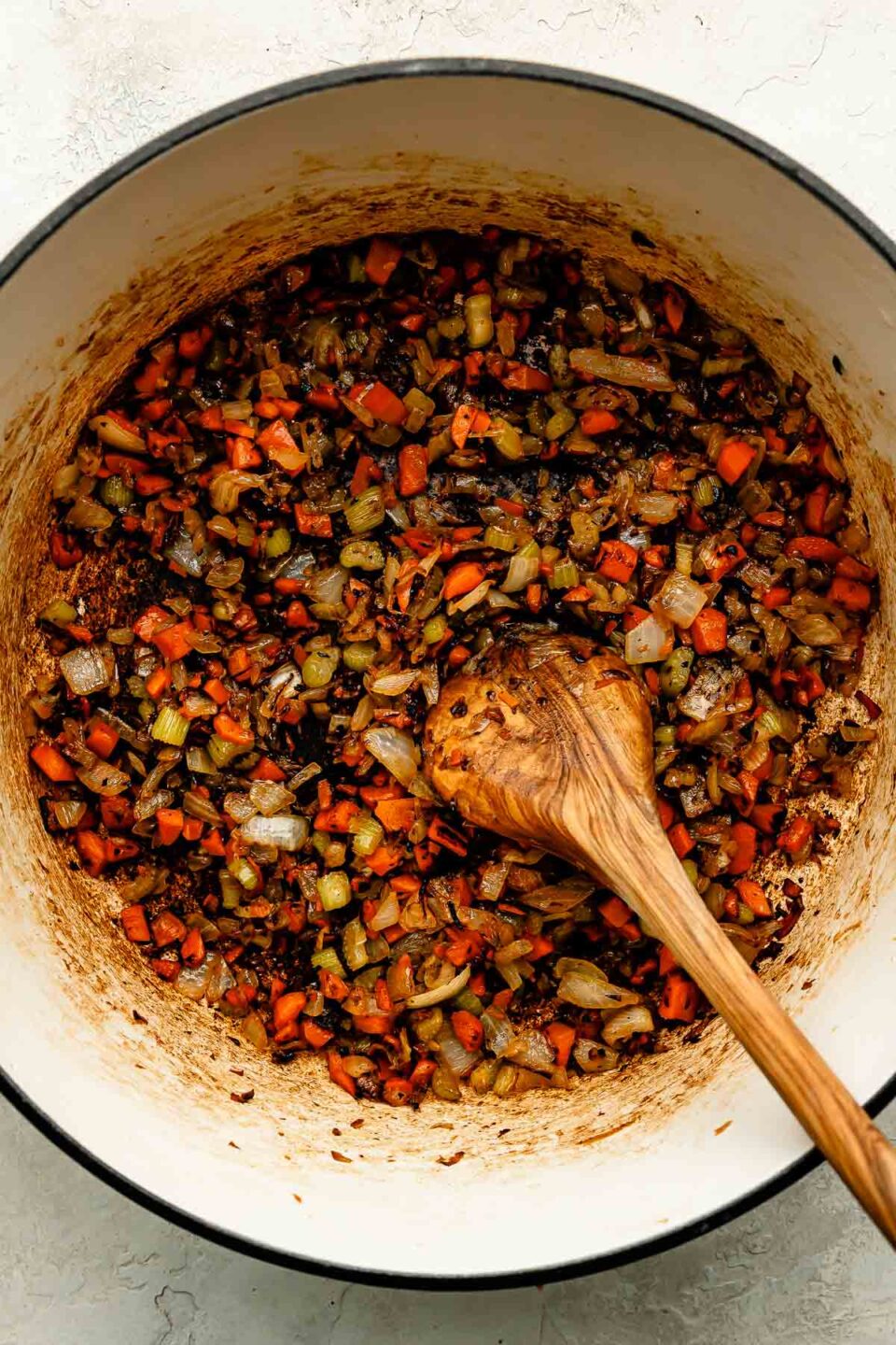 An overhead shot of browned soffritto with a wooden spoon in a Dutch oven atop an off-white surface.