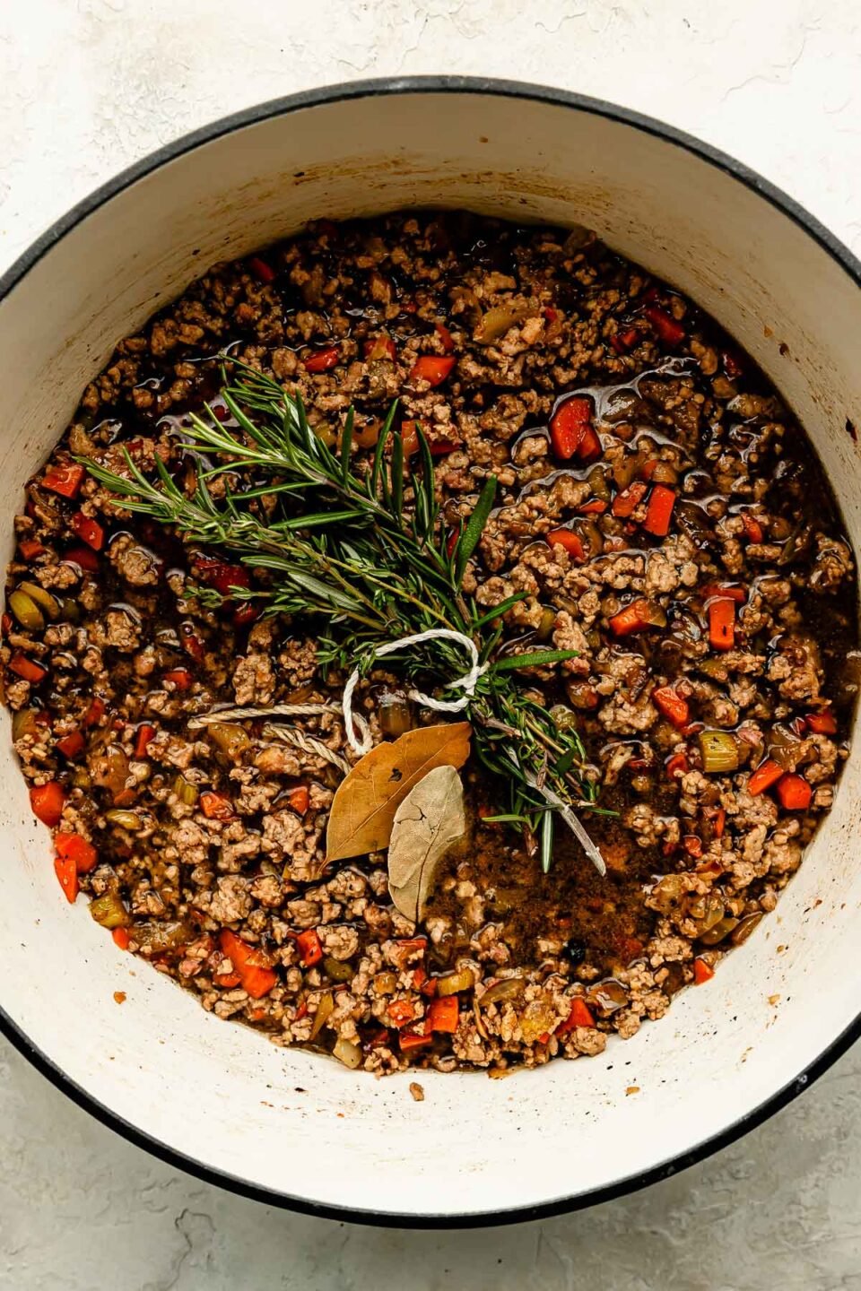 An overhead shot of pancetta, soffritto, ground pork, white wine, fresh herbs and bay leaves in a Dutch oven atop an off-white surface.