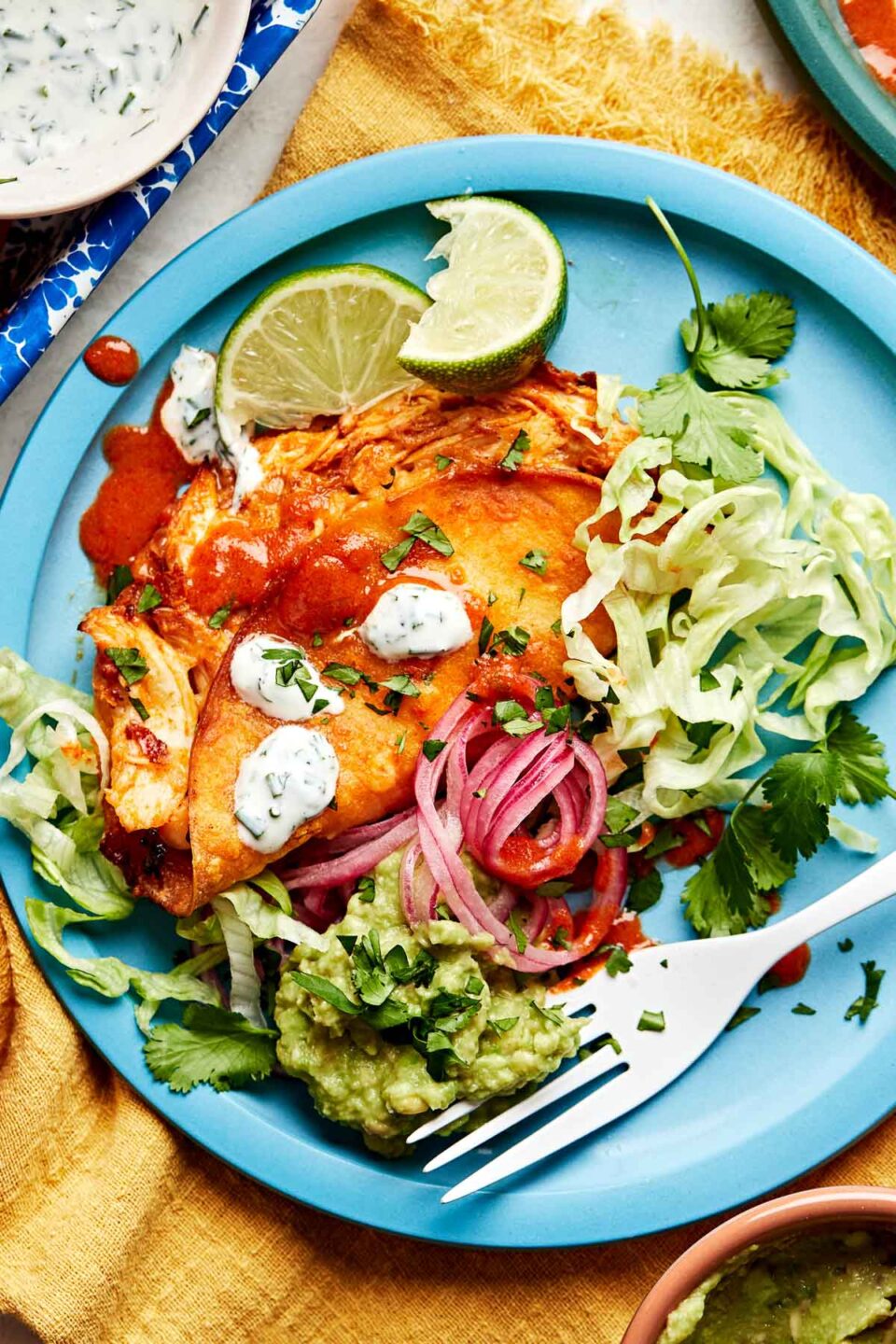 An overhead shot of a baked buffalo chicken taco garnished with ranch, buffalo sauce, limes and cilantro alongside pickled onions, guacamole, and shredded lettuce on a blue plate atop a yellow cloth.