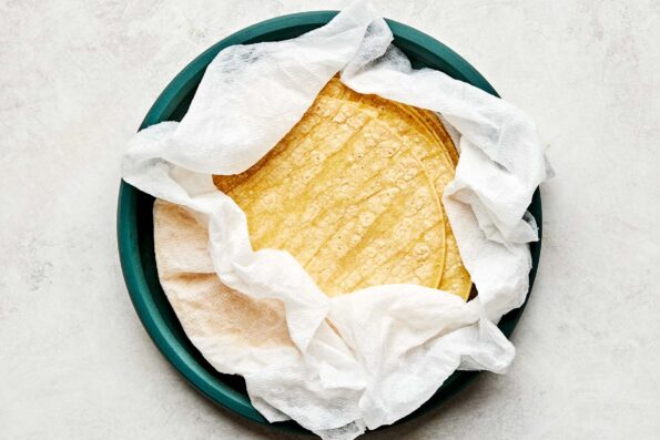 An overhead shot of corn tortillas wrapped in a damp paper towel on a teal plate atop a white marbled surface.