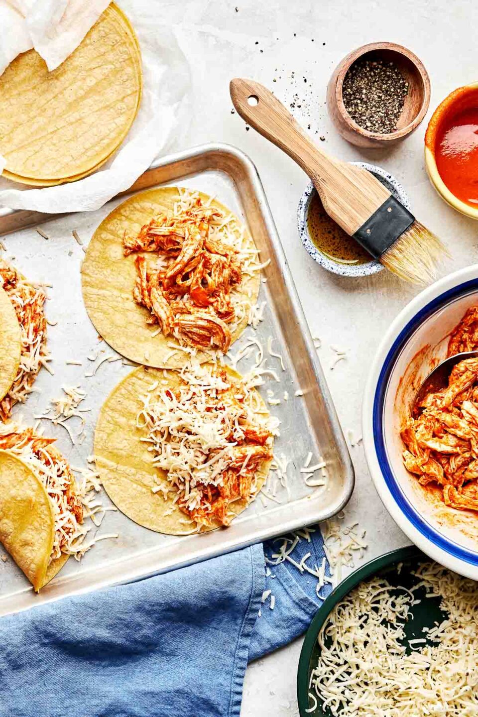 An overhead shot of two assembled and two partially assembled buffalo chicken tacos on a baking sheet atop a white surface. Corn tortillas and bowls of buffalo chicken, shredded cheese, olive oil, buffalo sauce and pepper sit alongside the sheet pan.