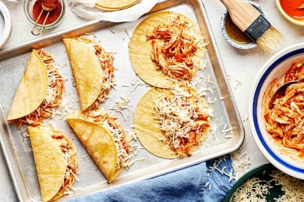 An overhead shot of two assembled and two partially assembled buffalo chicken tacos on a baking sheet atop a white surface. Corn tortillas and bowls of buffalo chicken, shredded cheese, olive oil, buffalo sauce and paprika sit alongside the sheet pan.
