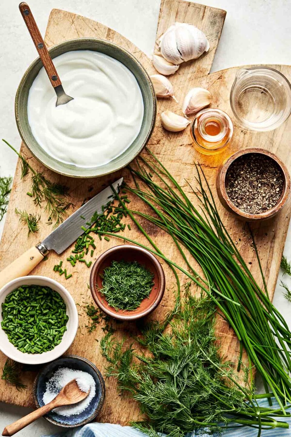 An overhead shot of ingredients displayed on a wooden cutting board stop a white surface: greek yogurt, garlic, chives, fresh dill, salt, pepper, vinegar and water.