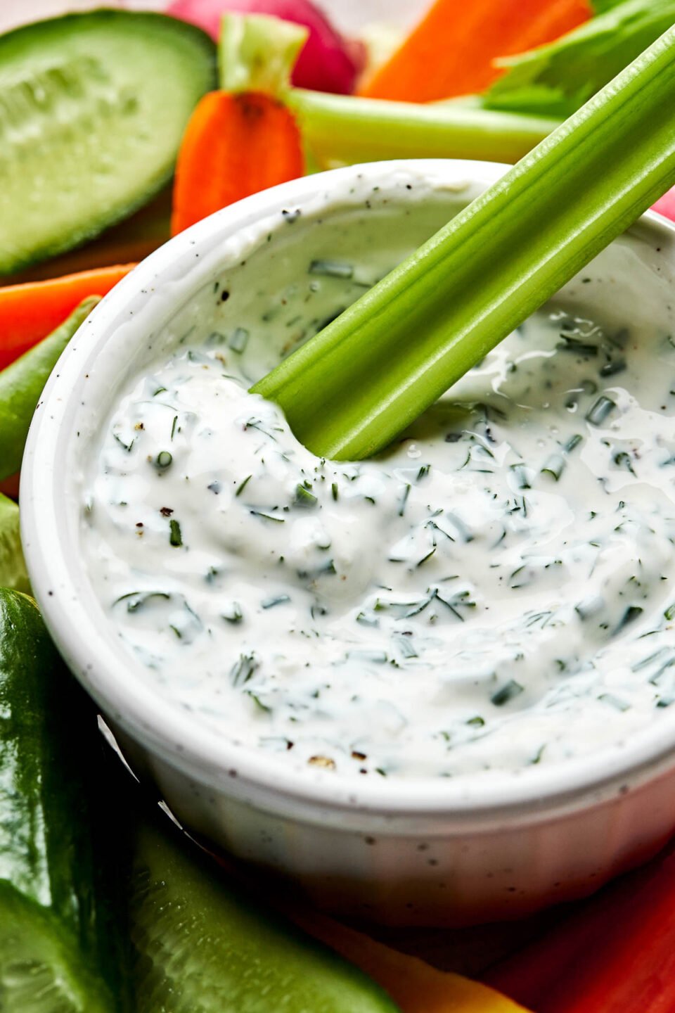 A close-up angled shot of a celery stick being dipped into a small white bowl of greek yogurt ranch, alongside sliced carrots, celery, radishes, snap peas and cucumbers.