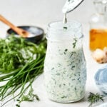 A side shot of a spoon dripping greek yogurt ranch into a glass jar full of ranch dressing. The jar sits on a white surface alongside a blue and white striped cloth, a container of vinegar, garlic, dill, chives, and a bowl of salt.