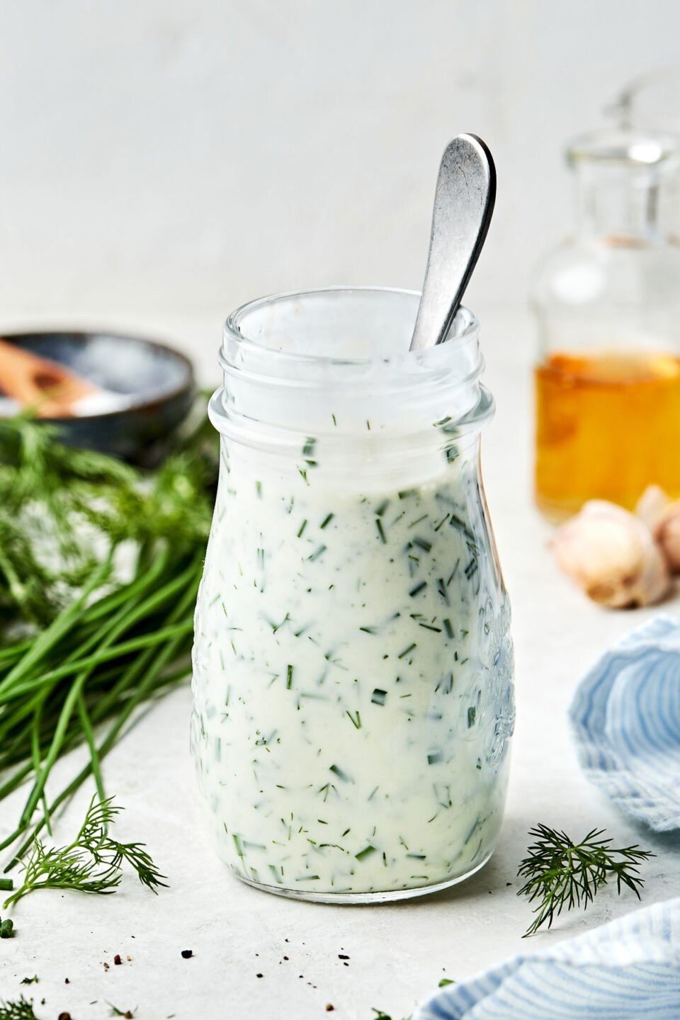 A side shot of ranch dressing in a glass jar. The jar sits on a white surface alongside a blue and white striped cloth, a container of vinegar, garlic, dill, chives, and a bowl of salt.