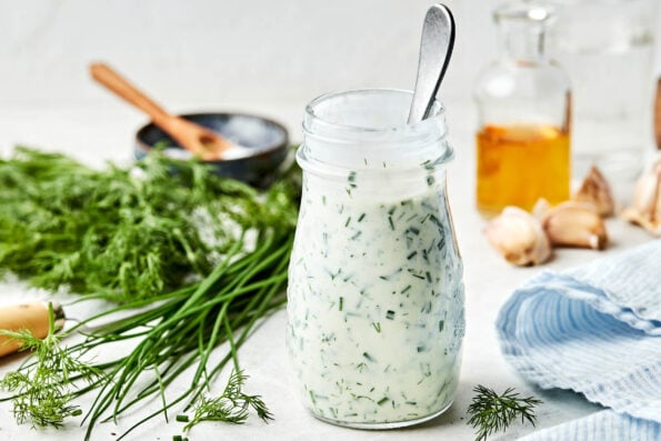 A side shot of greek yogurt ranch in a glass jar. The jar sits on a white surface alongside a blue and white striped cloth, a container of vinegar, garlic, dill, chives, and a bowl of salt.