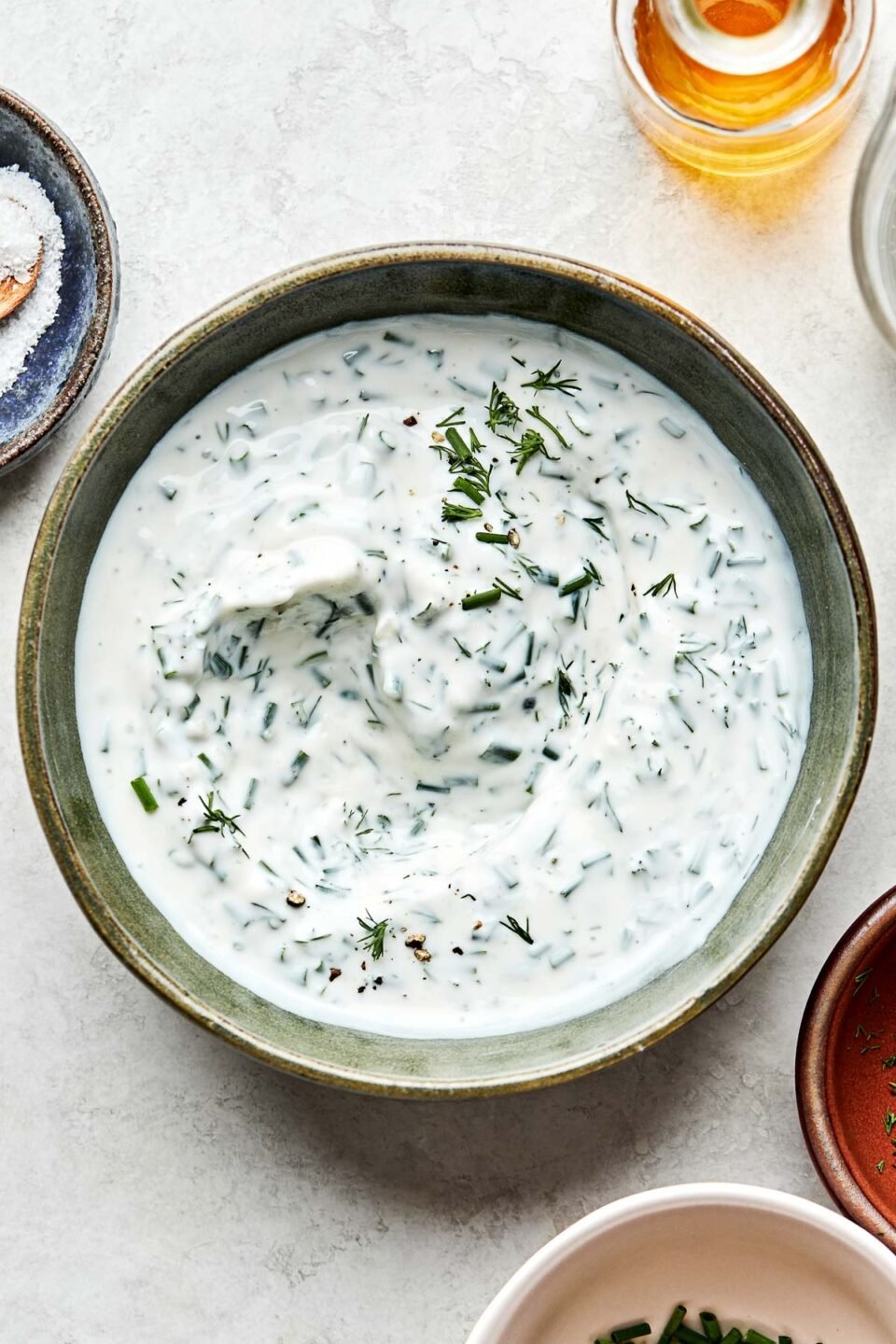 An overhead shot of ranch dip in a green stoneware bowl stop a white marbled surface. Dishes of vinegar, chives, dill, and salt sit alongside the bowl.