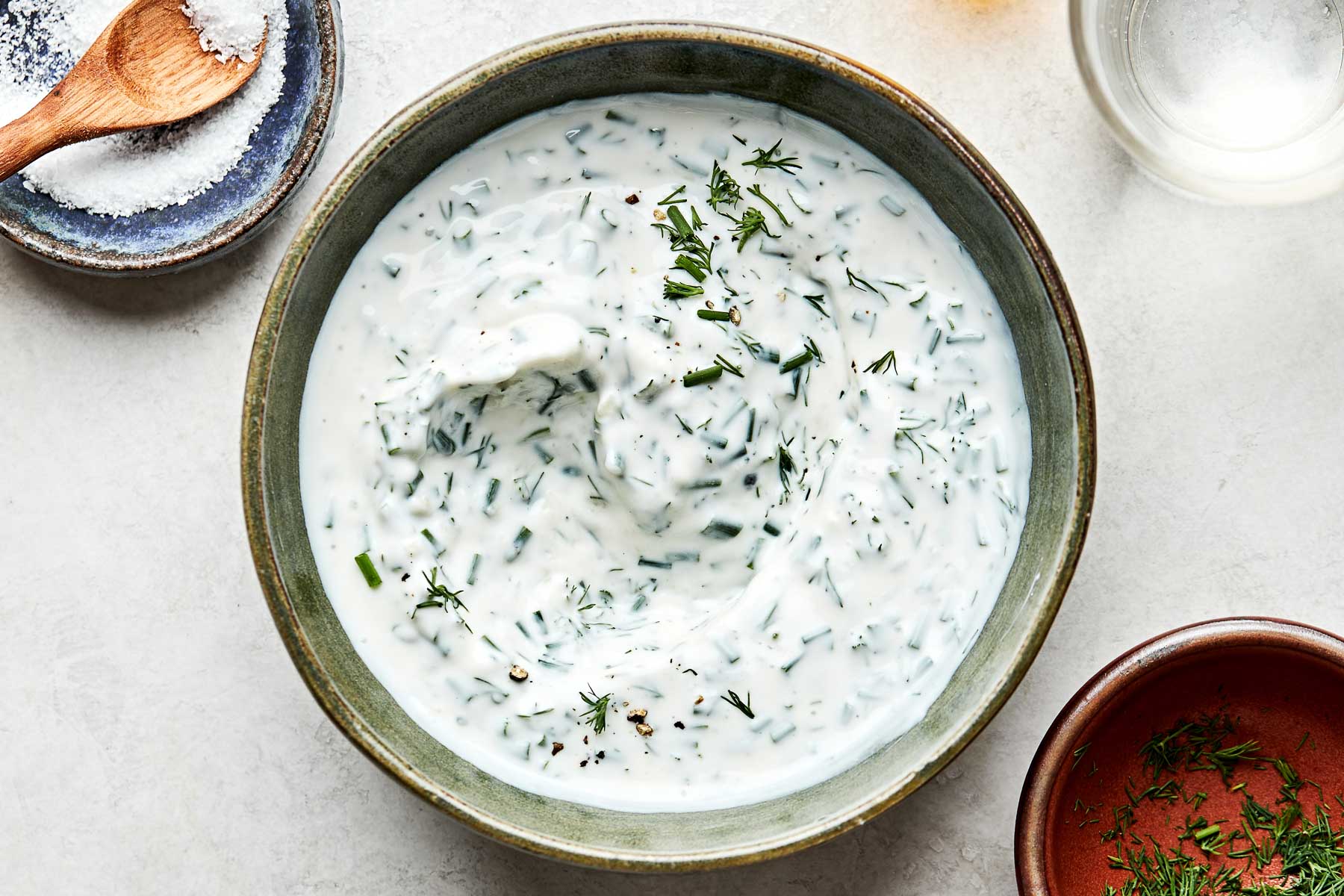 An overhead shot of greek yogurt ranch dressing in a green stoneware bowl stop a white marbled surface. Dishes of water, dill, and salt sit alongside the bowl.