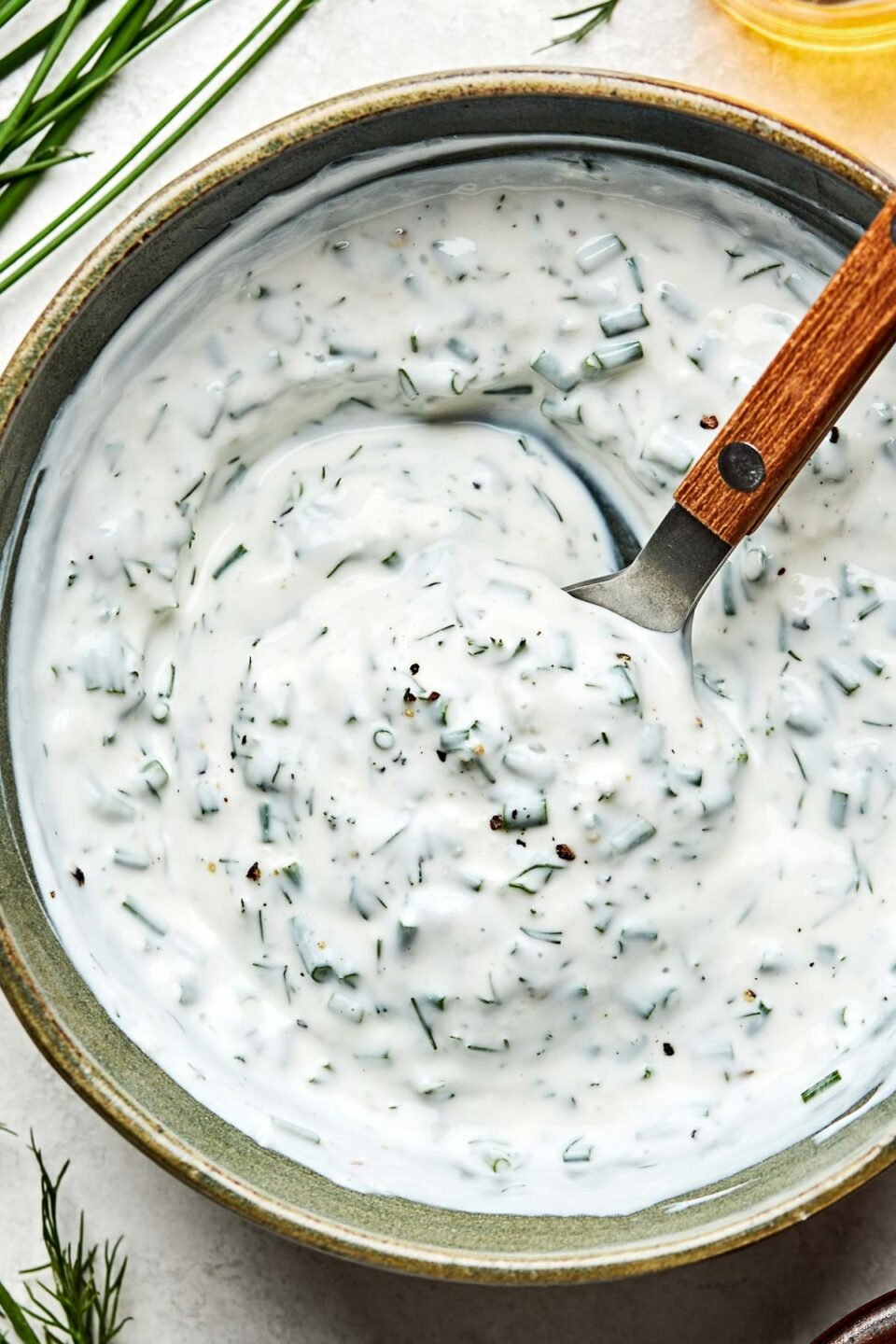 A close-up overhead shot of a bowl of yogurt ranch dressing with a spoon in it atop a white marbled surface. Fresh chives and dill sit alongside the bowl.