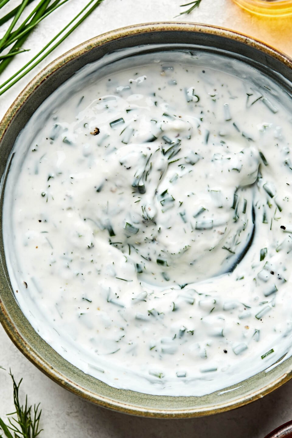 A close-up overhead shot of a bowl of ranch dip atop a white marbled surface. Fresh chives and dill sit alongside the bowl.
