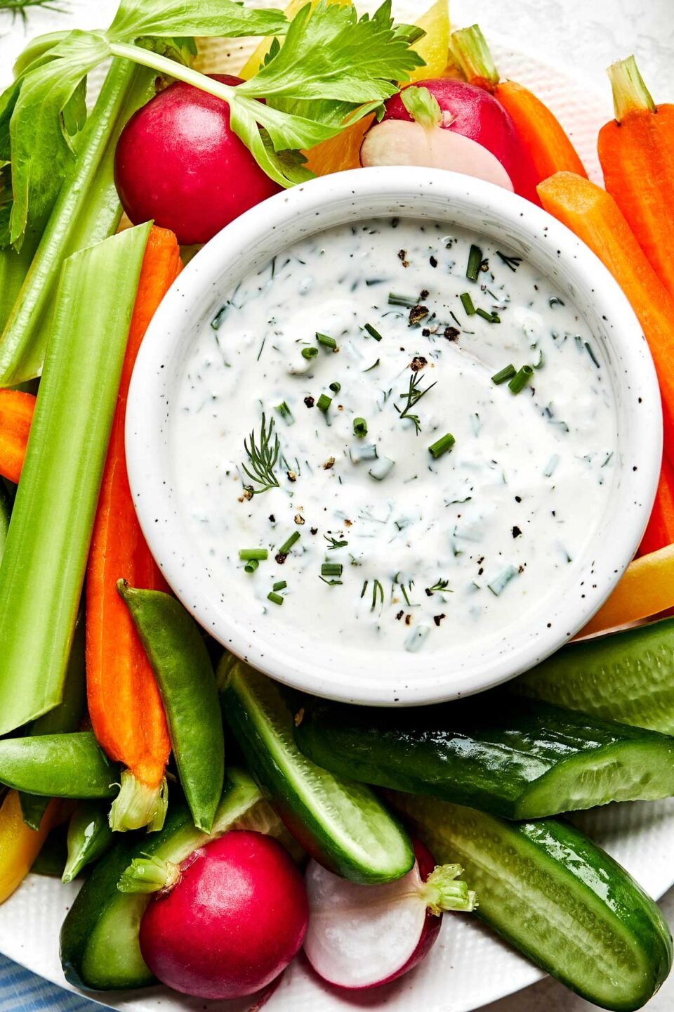 An overhead shot of a small white bowl of ranch dip garnished with fresh chives and dill alongside sliced carrots, celery, radishes, snap peas and cucumbers on a white plate.