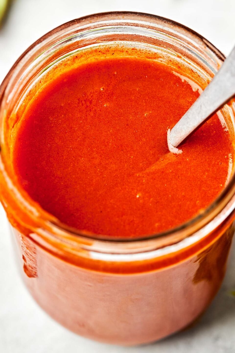 A close-up angled overhead shot of a jar of buffalo sauce with a spoon in it atop a white marbled surface.