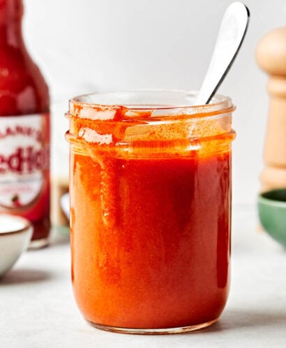 A side shot of a glass jar of homemade buffalo sauce sitting on a white surface alongside a bottle of Frank's Red Hot, small bowls, and a pepper grinder.