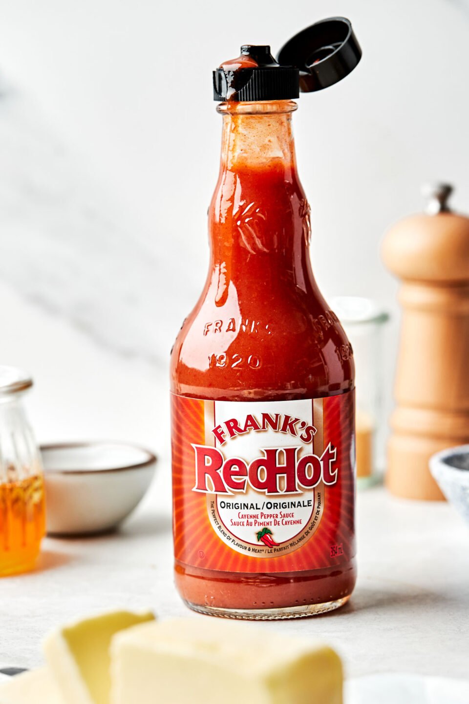 A side shot of a glass bottle of Franks Red Hot on a white surface in front of a white background.