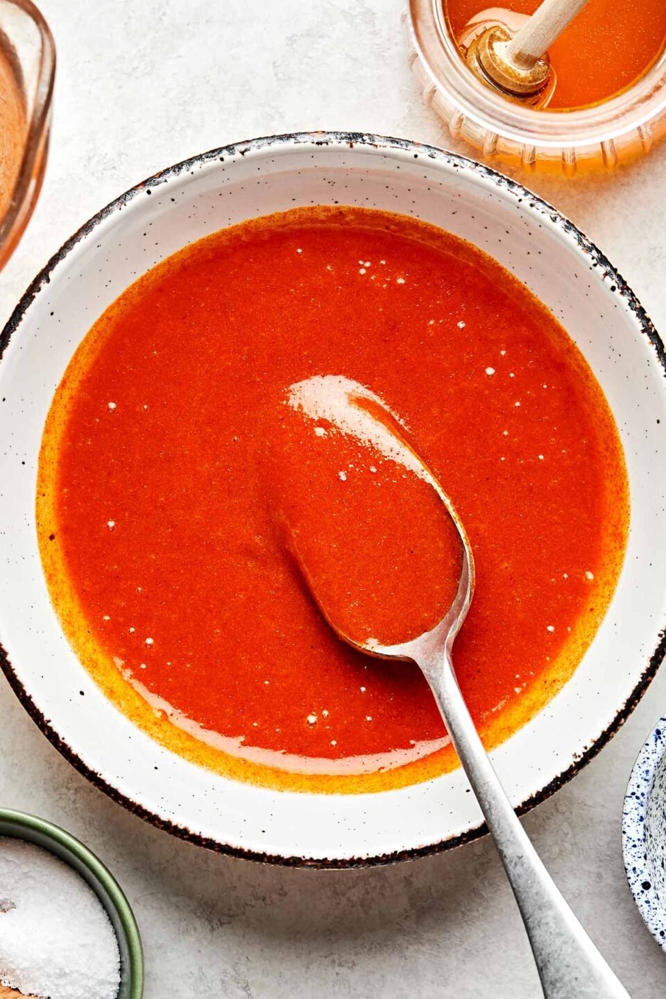 An overhead shot of buffalo sauce being held up with a spoon in a white stoneware bowl atop a white marbled surface.