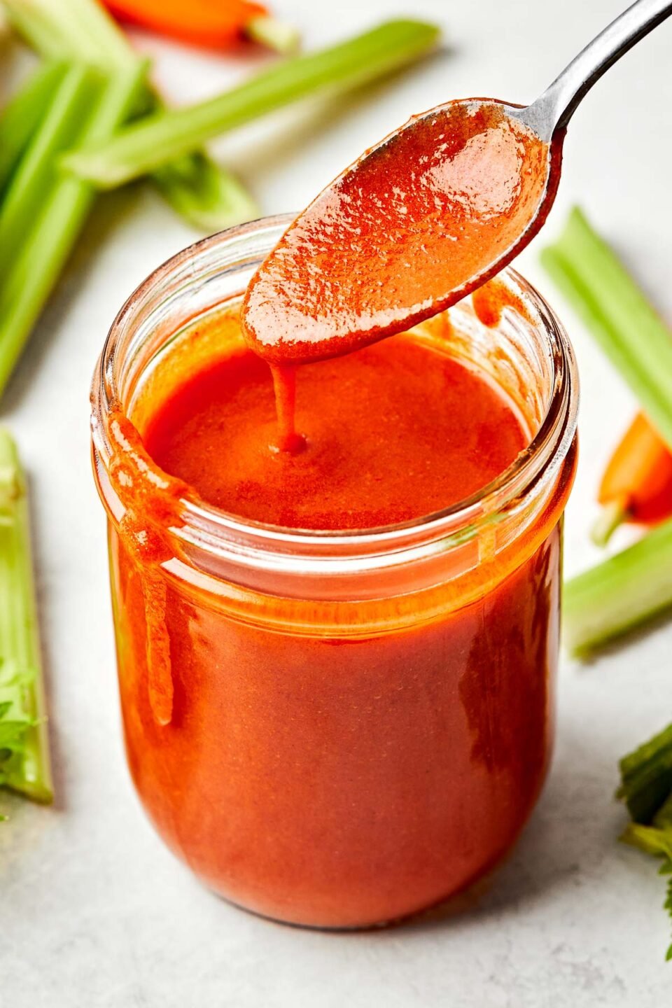 A side-angle shot of a spoon holding up buffalo sauce from a glass jar sitting atop a white marbled surface. Carrot and celery sticks sit alongside the jar.