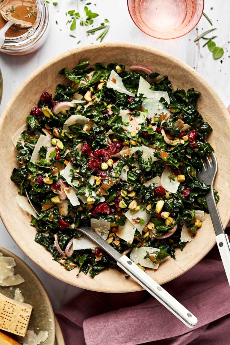 An overhead shot of kale salad in a large wooden bowl atop a white surface. A bowl of parmesan, a pink glass of water, a pink cloth and a jar of vinaigrette sit alongside the bowl.