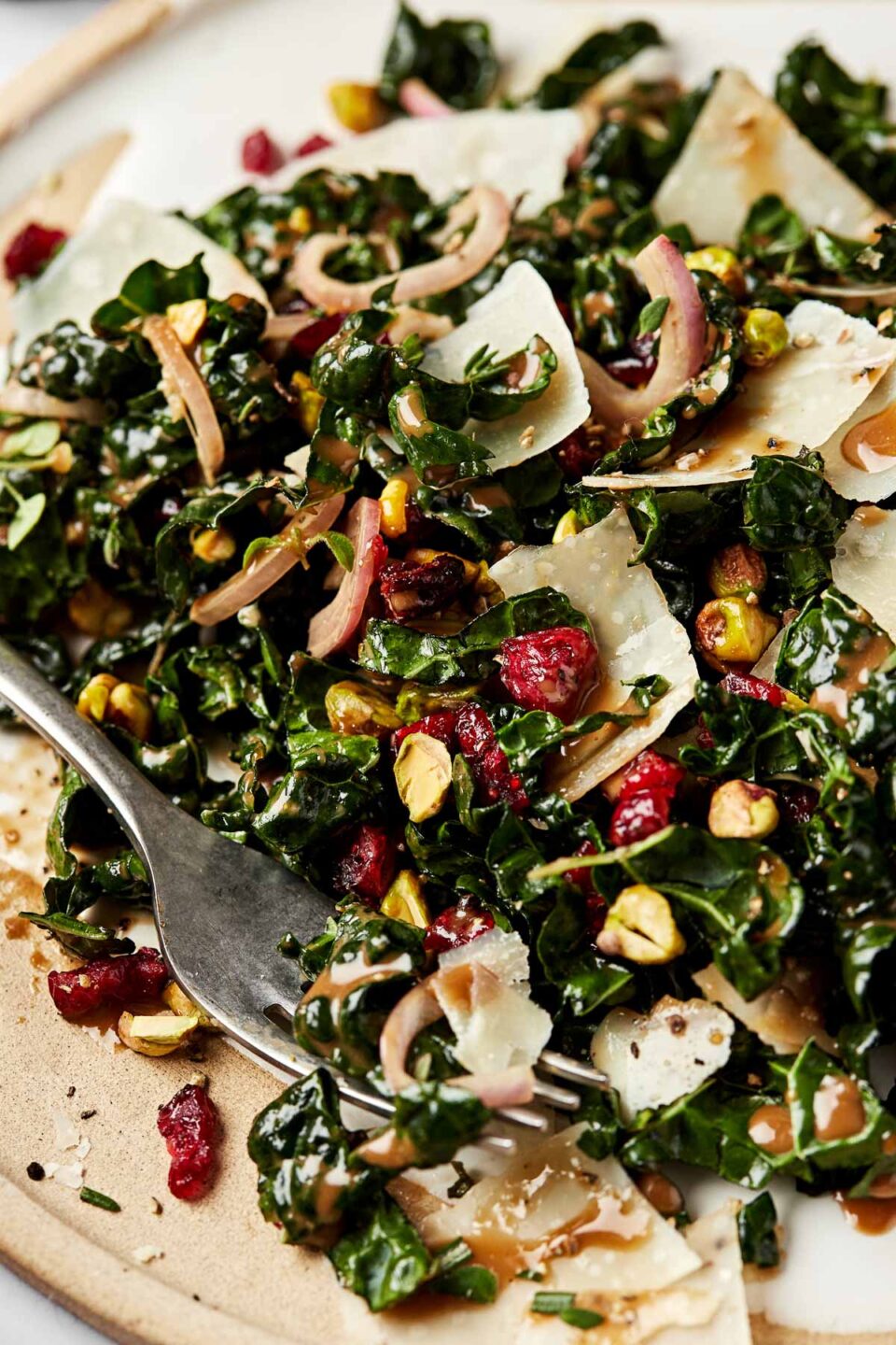 A closeup shot of a kale salad with cranberries, pistachios and parmesan on a beige plate with a silver fork.