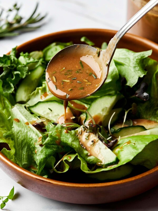 A spoon drizzles maple balsamic vinaigrette onto a salad in a large wooden bowl atop a white marbled surface. A jar of dressing can be seen in the background.
