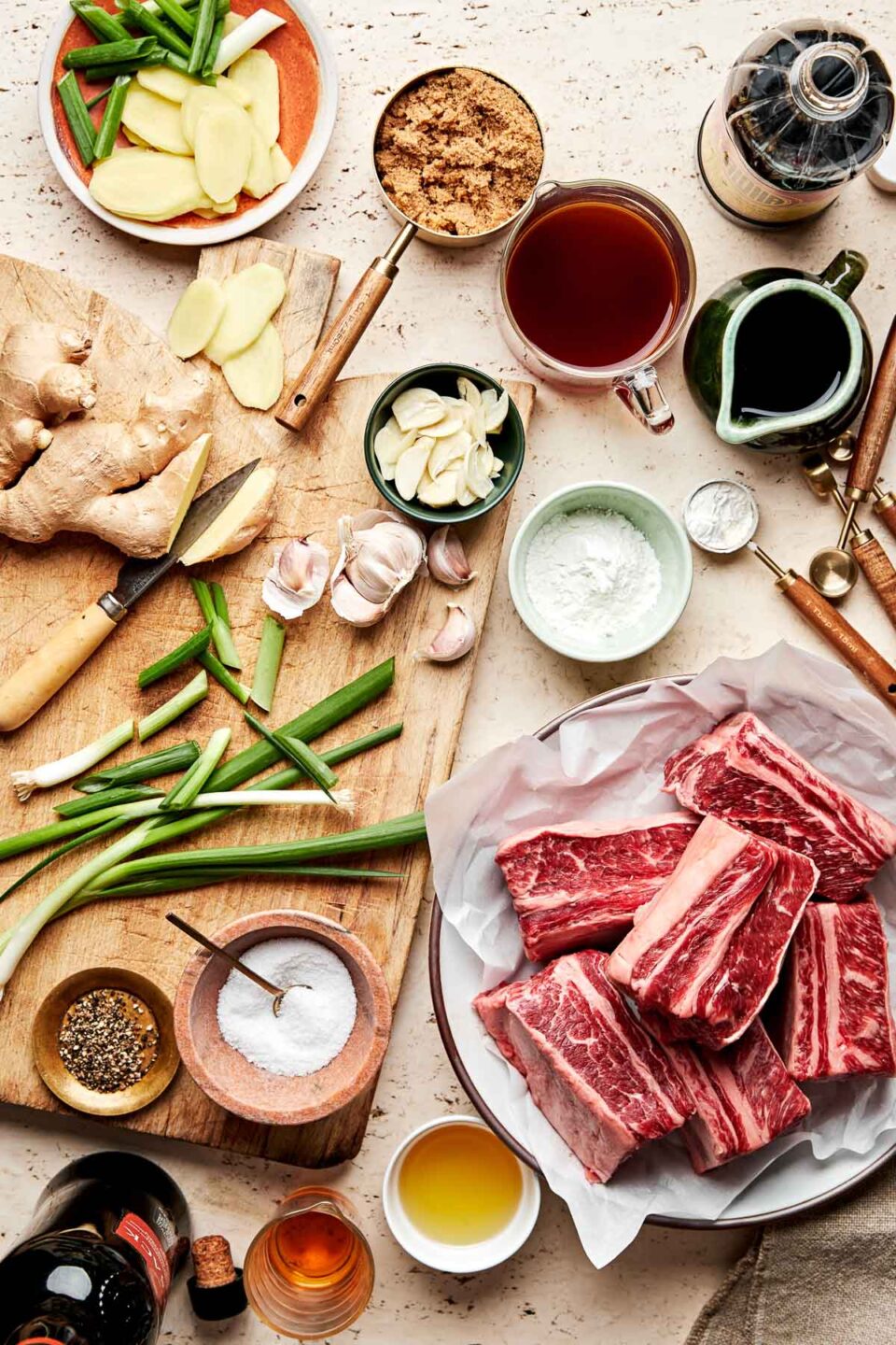 An overhead shot of ingredients displayed on a wooden cutting board and an off-white surface: short ribs, salt and pepper, vegetable oil, ginger, garlic, green onions, brown sugar, shoyu, beef broth, dry sherry, and cornstarch.