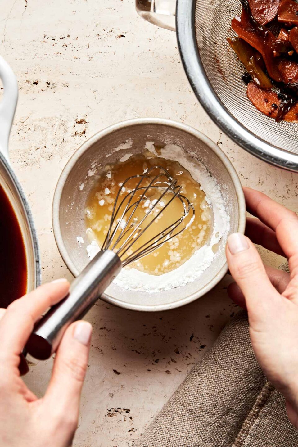 An overhead shot of a woman's hands mixing a cornstarch slurry in a beige stoneware bowl atop a textured beige surface. A strainer with strained aromatics and the pot of braising liquid sit alongside the bowl.