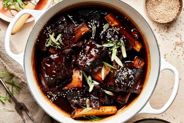 An overhead shot of soy-braised short ribs in a thickened braising liquid in a white pot atop a beige surface. A plate of sliced green onions and a bowl of sesame seeds sit alongside the pot.