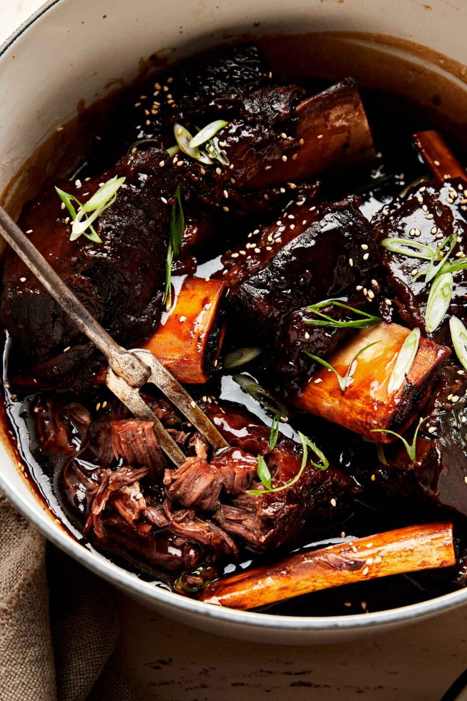A close-up shot of a two-pronged serving fork shredding a soy-braised short rib in a white pot full of braised ribs and thickened braising liquid garnished with green onions and sesame seeds.