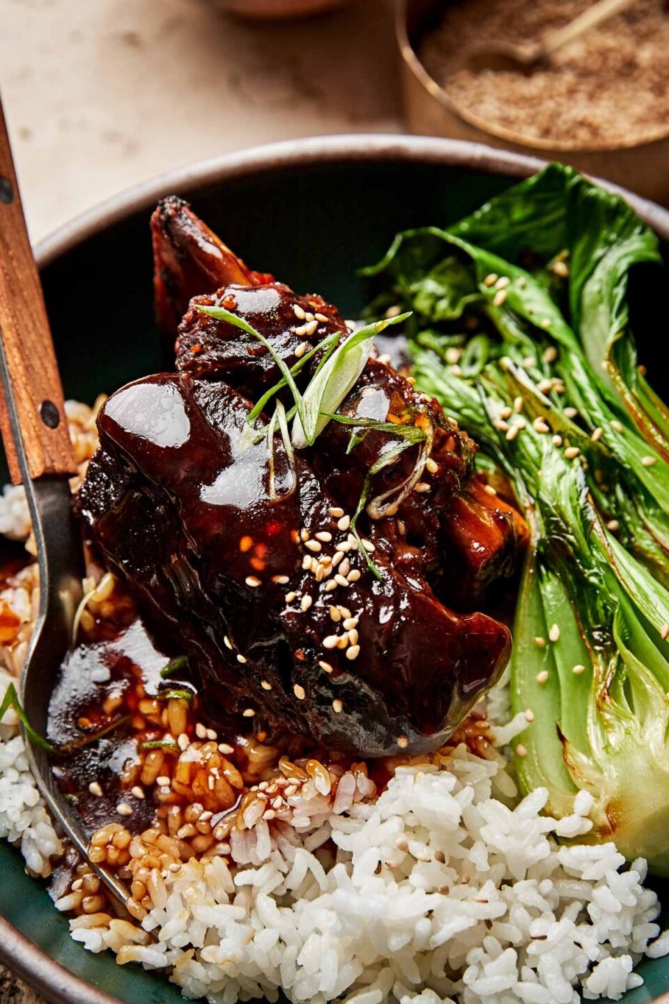 A close-up shot of a soy-braised short rib topped with thickened braising liquid in a dark green bowl atop a bed of white rice and steamed bok choy.