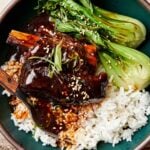 A close-up overhead shot of a soy-braised short rib topped with thickened braising liquid in a dark green bowl atop a bed of white rice and steamed bok choy.