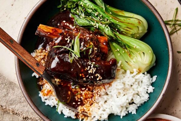 A close-up overhead shot of a soy-braised short rib topped with thickened braising liquid in a dark green bowl atop a bed of white rice and steamed bok choy.