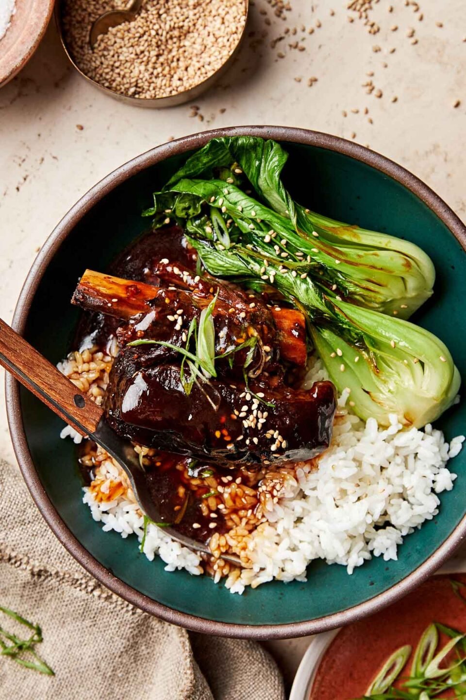 A close-up overhead shot of a soy-braised short rib topped with thickened braising liquid in a dark green bowl atop a bed of white rice and steamed bok choy. The bowl sits atop a beige surface alongside a dish of green onions and a bowl of sesame seeds.