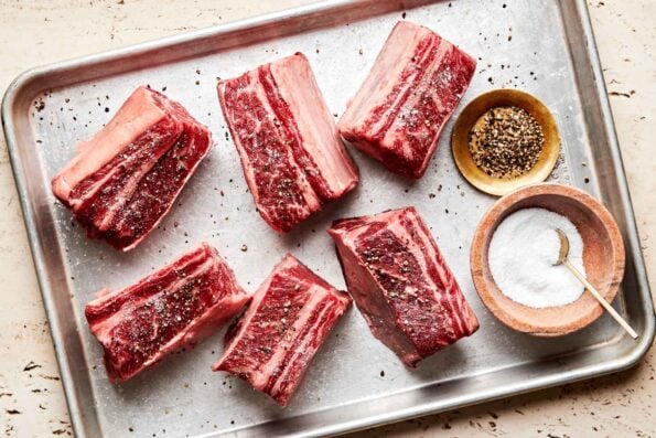 An overhead shot of six English-cut beef short ribs, generously seasoned with salt and pepper, alongside small bowls of salt and pepper on a sheet pan atop an off-white surface.