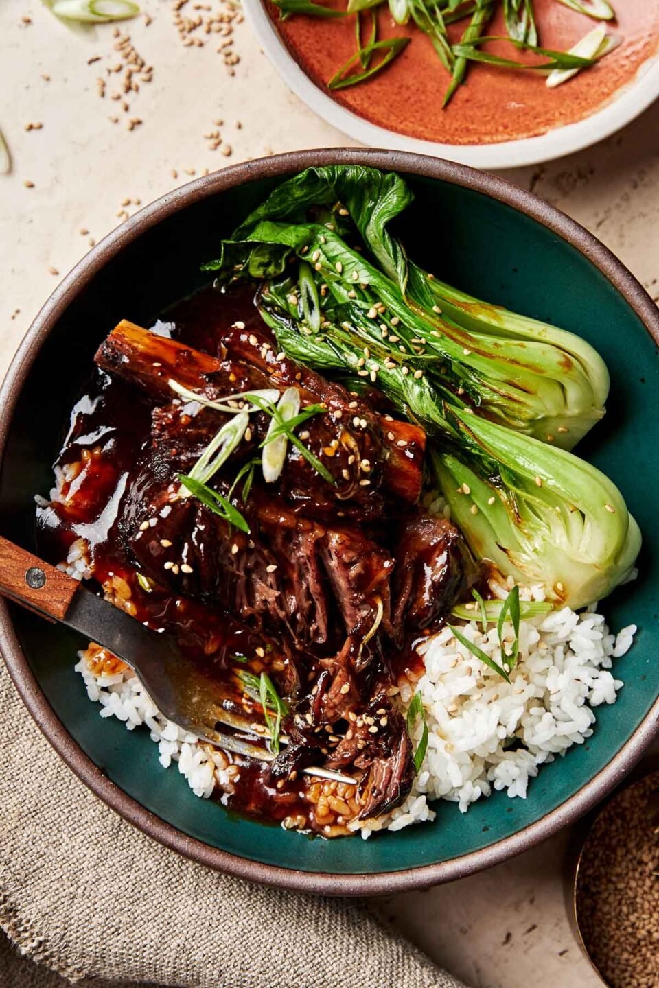 A close-up overhead shot of a soy-braised short rib topped with thickened braising liquid in a dark green bowl atop a bed of white rice and steamed bok choy. The bowl sits atop a beige surface alongside a dish of green onions and a bowl of sesame seeds.