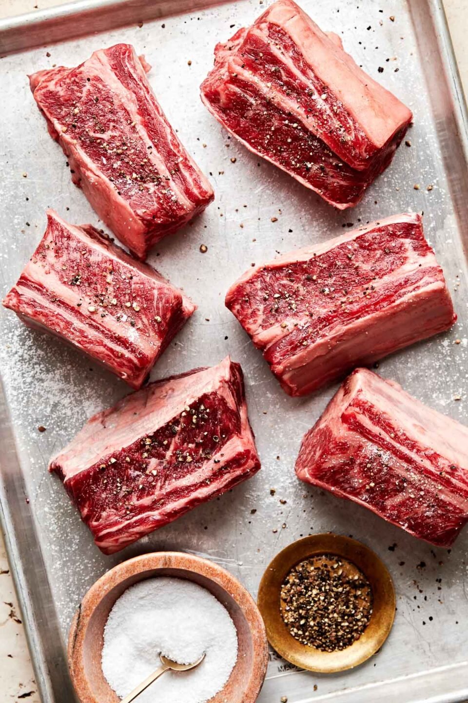 An overhead shot of six English-cut beef short ribs, generously seasoned with salt and pepper, alongside small bowls of salt and pepper on a sheet pan atop an off-white surface.