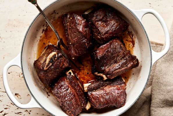 An overhead shot of six browned short ribs in a white pot atop an off-white surface. A silver two-pronged fork sits in the pot.