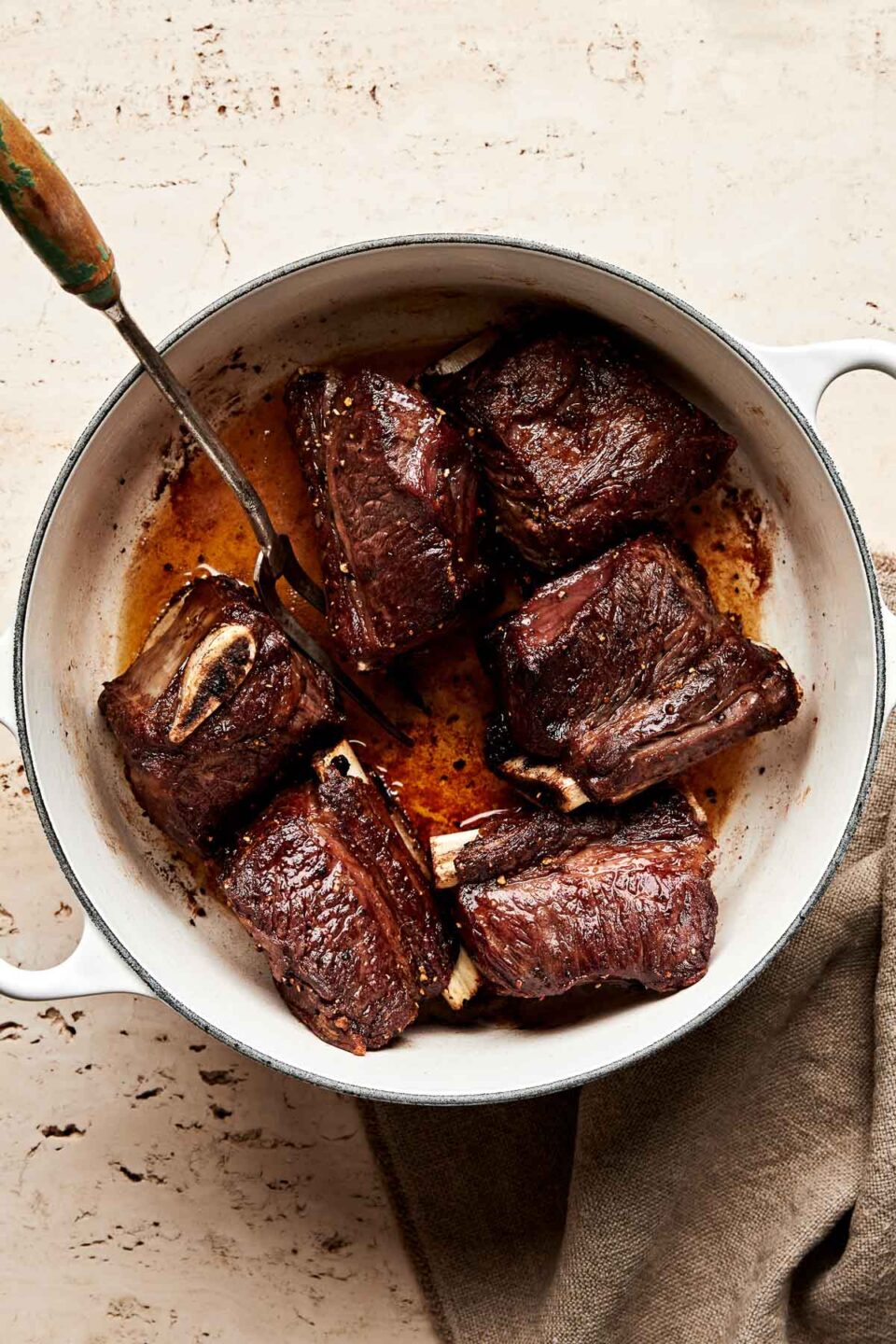 An overhead shot of six browned short ribs in a white pot atop an off-white surface. A silver two-pronged fork sits in the pot.