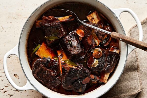 An overhead shot of soy-braised short ribs in a large white pot atop an off-white surface. A large wood-handled spoon rests in the pot.