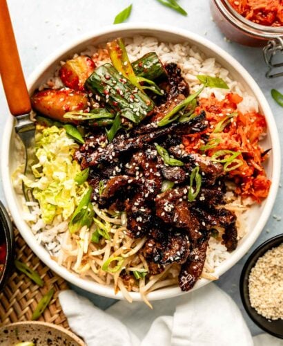 An overhead shot of sliced bulgogi steak in a bowl with cabbage, cucumber kimchi, and white rice. A bowl of sesame seeds and a jars of kimchi, sliced green onions and cucumber kimchi sit alongside it.