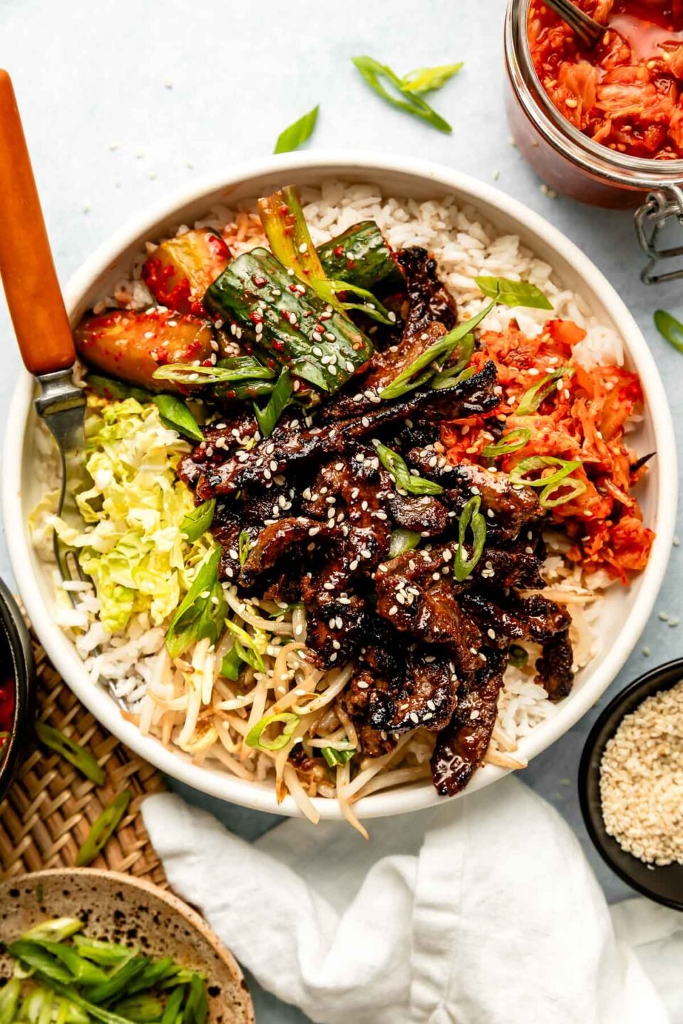 An overhead shot of sliced bulgogi steak in a bowl with cabbage, cucumber kimchi, and white rice. A bowl of sesame seeds and a jars of kimchi, sliced green onions and cucumber kimchi sit alongside it.