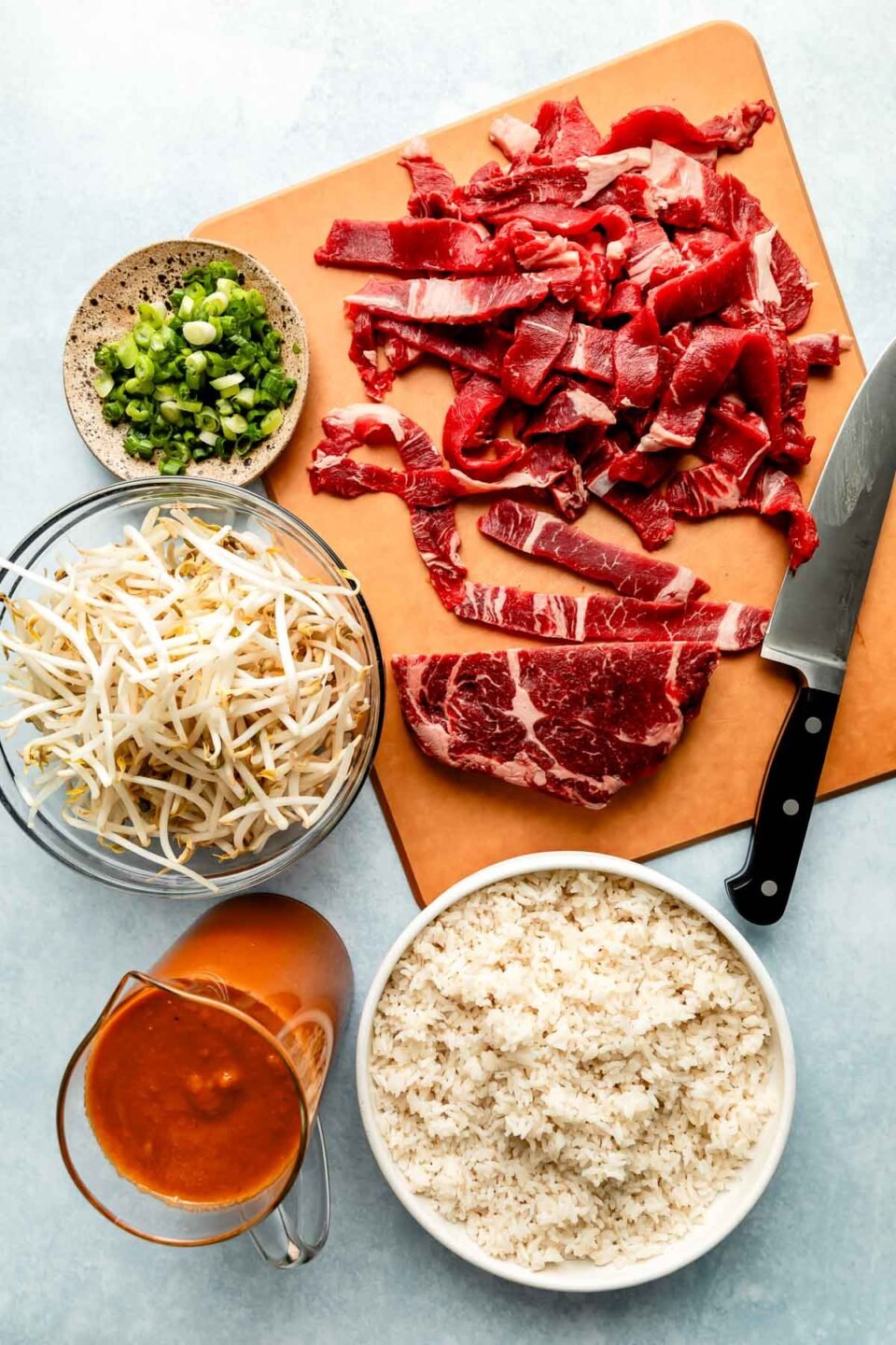 An overhead shot of ingredients in bowls and on a cutting board on a grey surface: sliced ribeye steak, bulgogi marinade, mung bean sprouts, green onions, and white rice.