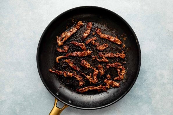 An overhead shot of thin strips of marinated steak being seared in a black nonstick skillet atop a grey surface.