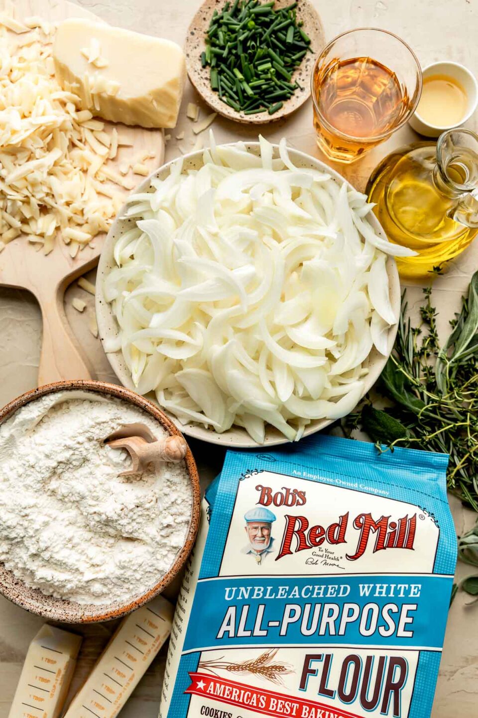 An overhead shot of ingredients displayed in bowls and on a wooden board atop a beige surface: an unopened bag of Bob's Red Mill flour, a bowl of flour, sliced onions, gruyere cheese, chives, fresh herbs, butter, olive oil, sherry, and apple cider vinegar.