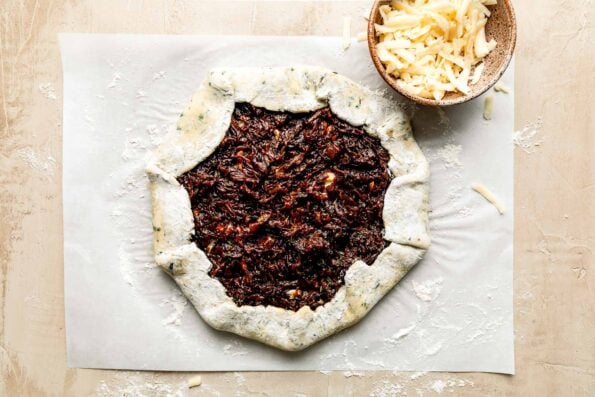 An overhead shot of an assembled French onion galette on a piece of parchment paper atop an off-white surface. A small bowl of shredded gruyere cheese sits alongside it.